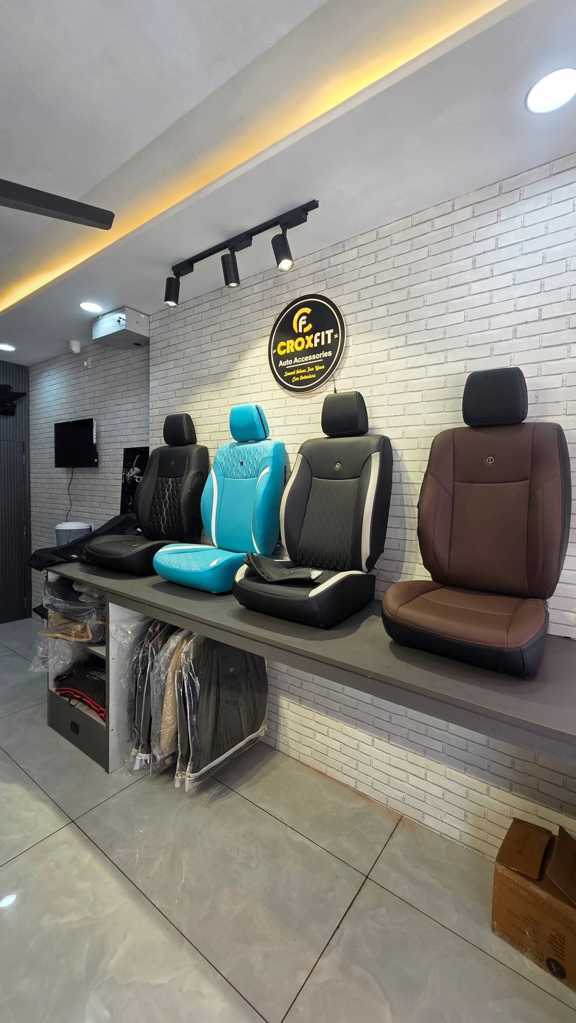 Car seat cover display on a grey brick wall lit by black track lights, Malik Electronics showroom, Ludhiana