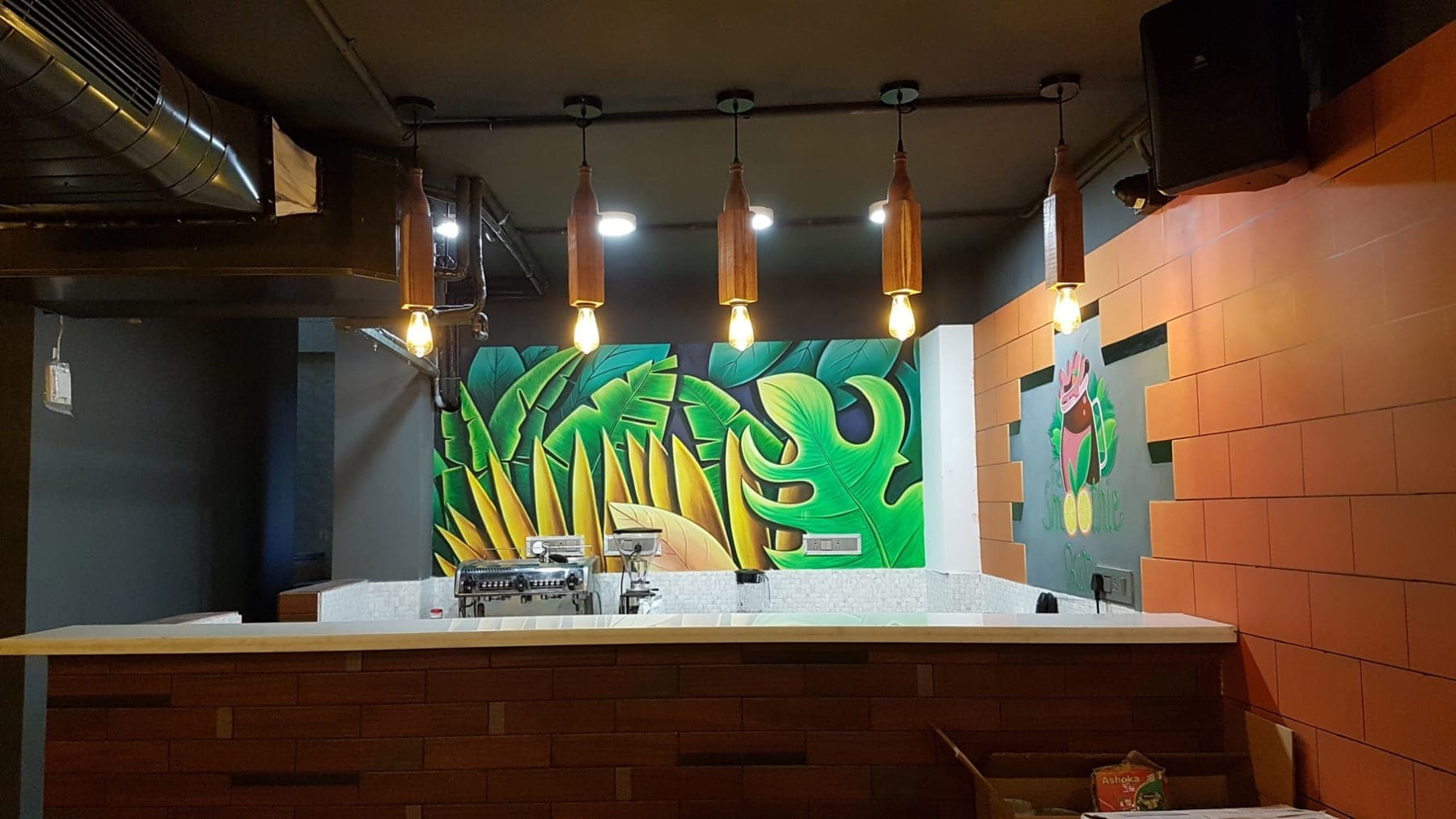 Tropical leaf mural behind the juice bar counter with wooden bottle pendant lights, FiTreat restaurant, Ludhiana