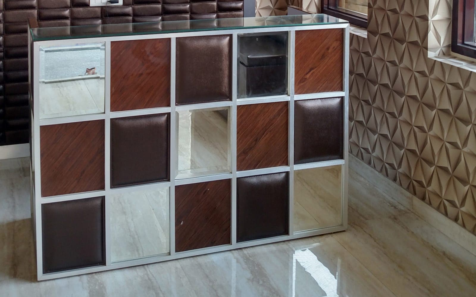Mirrored checkerboard reception desk with wood-grain and leather panels in a chrome frame, beside geometric 3D wallpaper, Ludhiana