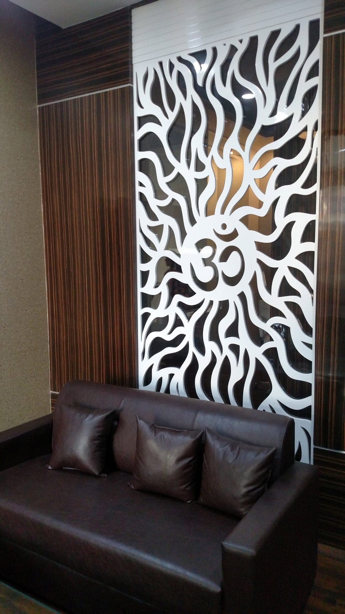 White CNC-cut Om jaali partition against wood-grain laminate paneling with a dark leather sofa, Ludhiana office