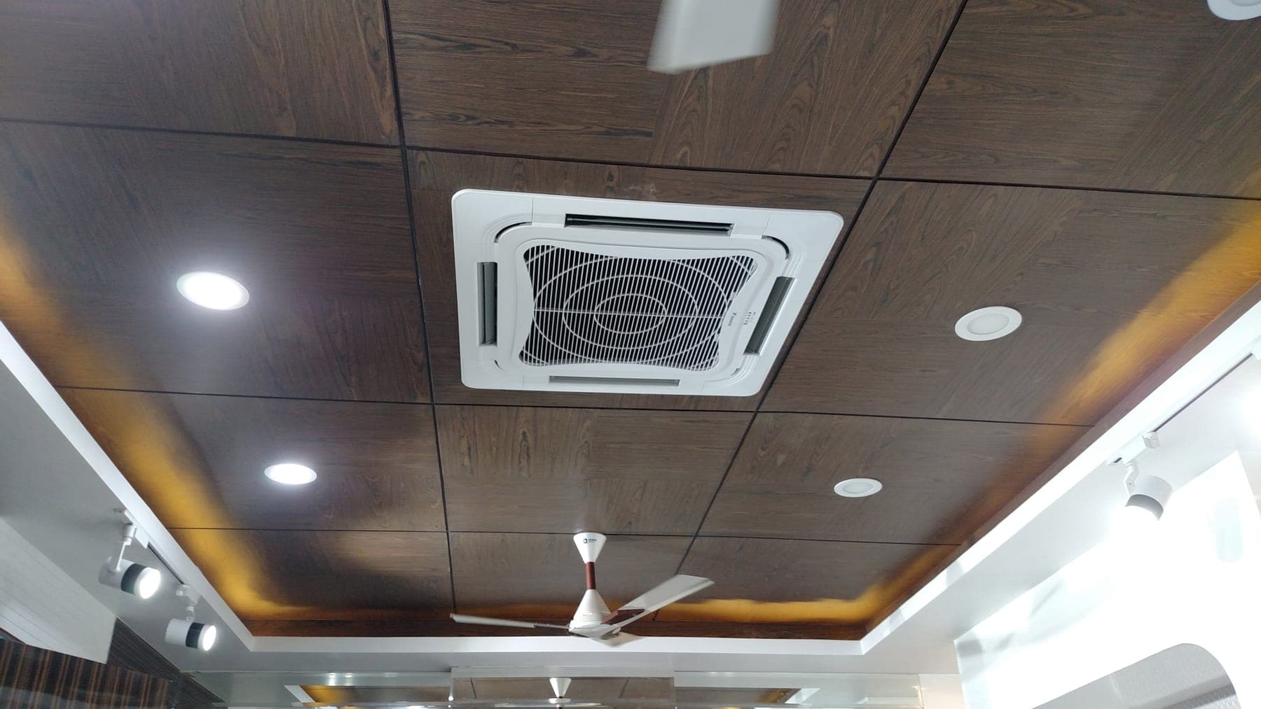 Dark wood-paneled ceiling with warm indirect LED cove lighting and white track lights, Ludhiana office interior